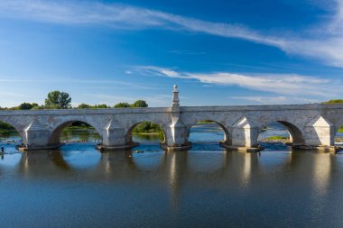 Fransa 'nın orta kesimindeki La Charite sur Loire' da Loire Nehri 'ni geçen tarihi bir kemer köprüsünün klasik görüntüsü. Sahnede sakin su, mavi gökyüzü ve yumuşak gün ışığı altında yemyeşil nehir kıyıları yer alıyor..