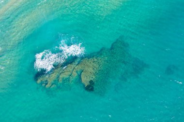 Yüzeyinde hafif dalgalar kırılan berrak turkuaz suyun altındaki su altındaki su altı kaya oluşumunun yukarıdan görünüşü. Güneş ışığı sualtı dokularını vurgular, canlı ve ferahlatıcı bir kıyı manzarası yaratır..