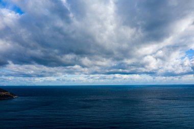 Geniş mavi deniz, kalın ve dokulu bulutlarla dolu bir gökyüzünün altındaki ufka kadar uzanıyor. Dengesiz atmosfer, değişen ışık ve suyun üzerindeki derin gölgelerle güçlenir..