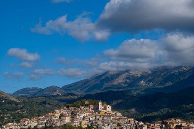 İtalya 'nın Rivello kentindeki tepe köyünde pastel renkli evler ve engebeli dağlara karşı kurulu önemli bir kilise ve dramatik bulutlarla dolu bir gökyüzü bulunuyor. Güneş ışığı binaların dokusunu ve bereketli manzarayı vurgular.