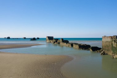 Tarihi bir yapay limanın beton kalıntıları sakin turkuaz sulara doğru süzülüyor, yumuşak kum ve berrak mavi gökyüzü Asnelles, Normandiya yakınlarında sakin bir kıyı manzarası yaratıyor..