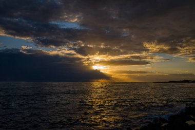 Dramatic golden sunlight breaks through dense clouds above the Adriatic Sea, casting vivid reflections across the rippling water. The moody sky and shimmering ocean create a striking coastal seascape at dusk.