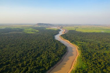 Kamboçya 'daki Tonle Sap yakınlarında sık orman ve yamalı tarım arazileri boyunca kıvrılan çamurlu bir nehrin geniş hava görüntüsü. Manzara, bereketli bitki örtüsü, açık araziler ve berrak mavi gökyüzünün altındaki uzak tepeleri kapsar.