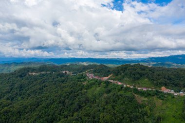 Geniş yeşil tepeler ve katmanlı bulutlarla dolu dramatik bir gökyüzünün altındaki uzak mavi dağlarla çevrili yemyeşil bir orman sırtına tünemiş bir köyün havadan görünüşü. Manzara uzak sükuneti ve doğal hatları yakalar.