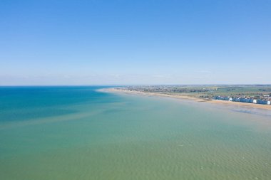 Courseulles-sur-Mer 'deki Juno Sahili' nin havadan görünüşü, durgun turkuaz sular, geniş kumlu sahil şeridi ve berrak mavi gökyüzü altında sıra sıra sahil kulübeleri..