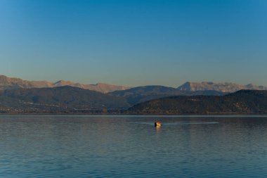 Tek bir tekne sessizce Ioannina Gölü 'nün sakin yüzeyinde süzülür. Uzak Pindus dağları ve sakin bir zemin oluşturan yumuşak yamaçlar. Yumuşak akşam ışığı ve açık gökyüzü Yunanistan 'ın Epirus kentinde sakin bir atmosfer yaratıyor.