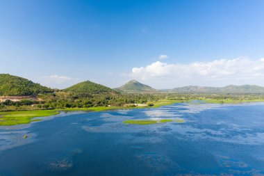 Brateak Krola Gölü 'nün geniş mavi suları Kamboçya' nın Kampong Chhnang kentindeki berrak bir gökyüzünün altında yemyeşil tepeler ve dağınık su bitkileri ile buluşur. Sahne, gün ışığında doğal dokuları ve açık alanı ön plana çıkarıyor..