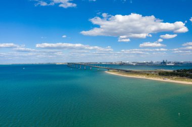 Pont de IIle de Re 'nin yüksek perspektifi anakaraya doğru canlı turkuaz suyu boyunca uzanır, kumlu kıyı şeridi ve dağınık kümülüs bulutları üzerimizde. Sahne açık kıyı manzarası ve sakin atmosferi vurgular