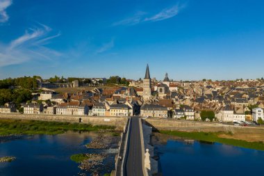 Loire nehri boyunca uzanan geniş bir taş köprü, berrak mavi gökyüzünün altında kendine özgü manastır kulesi ve kümelenmiş çatılarıyla La Charite sur Loire 'ın ortaçağ kalbine doğru uzanır. Nehir kenarı manzarası şehirlerin mimari mirasını ön plana çıkarıyor ve
