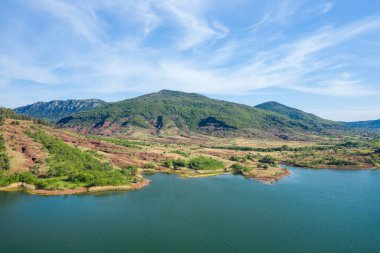 Lac de Salagou 'nun kızıl toprağın üzerinde yükselen yemyeşil tepeleri ve parlak, hafif bulutlu bir gökyüzünün altında beliren durgun mavi suları. Manzara canlı doğal renklere ve barışçıl, açık bir atmosfere sahiptir..