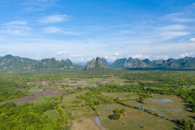 Yeşil pirinç tarlaları ve kırsal Laos kırsalındaki dağınık su havuzlarının üzerinde yükselen dramatik karst dağlarının geniş hava manzarası. Manzara bereketli bitki örtüsü, açık tarım arazisi ve yumuşak bulutlu canlı mavi bir gökyüzüne sahiptir..