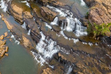 Hava perspektifi, güney Laos 'taki Tad Lo' daki katmanlı volkanik kayaların ve küçük çıkıntıların üzerinden akan suyu gösteriyor. Sahne, yumuşak gün ışığı altında zıt taş dokuları ve nazik çağlayanları vurguluyor..