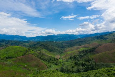 Kırsal Laos 'taki dağınık bulutlarla dolu geniş bir gökyüzünün altındaki uzak mavi dağ sırtlarına uzanan yuvarlanan yeşil tepelerin ve orman yamaçlarının geniş görüntüsü. Manzara doğal katmanlar, yemyeşil bitki örtüsü ve geniş bir his içerir.
