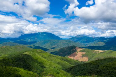 Yuvarlanan yeşil tepeler ve uzak dağ zirveleri, Phou Khoun ve Luang Prabang, Laos kırsallarındaki dramatik kümülüs bulutlarıyla dolu canlı mavi bir gökyüzünün altında uzanıyor. Manzara yemyeşil bitki örtüsü, açık yamaçlar ve zıtlık içerir.
