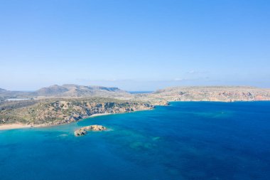 Girit 'teki Vai plajı yakınlarındaki derin mavi Akdeniz sularına giren engebeli bir kıyı şeridinin geniş hava manzarası. Sahnede güneşli uçurumlar, berrak turkuaz sığlıklar ve taze, davetkar bir atmosfer için parlak bir gökyüzü yer alıyor..