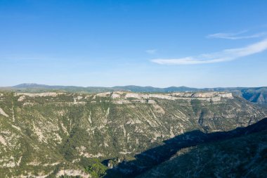 Navacelles Sirki 'ndeki derin bir orman kanyonunun üzerinde dramatik kireçtaşı uçurumları kulesi. Açık mavi gökyüzünün altındaki engebeli arazide güneş ışığı keskin gölgeler bırakıyor..