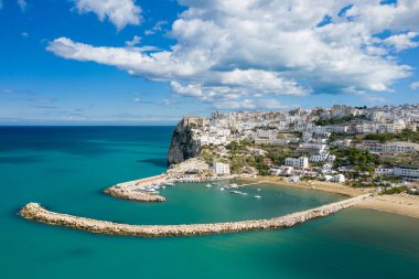 Peschici 'nin panoramik havası eğri taşlı dalgalı su üzerinde yükselen beyaz binalar ve dramatik bulutlarla dolu bir gökyüzünün altında berrak turkuaz su..