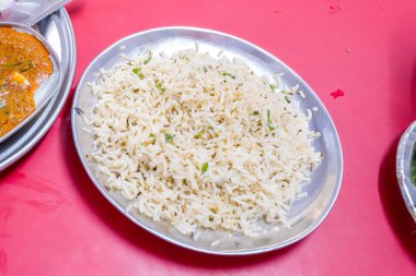 Paslanmaz çelik bir tabakta servis edilen otlarla süslenmiş tüylü basmati pirinci, Delhi 'deki sıradan bir restoranda canlı kırmızı bir masaya konmuş. Basit sunum tahılların dokusunu ve tazeliğini vurgular.