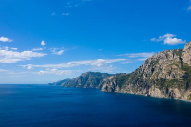 Engebeli kireçtaşı kayalıklarının, berrak bir gökyüzünün altında derin mavi Akdeniz sularıyla buluşması geniş bir manzara. Güneş ışığı, İtalyan kırsalının tipik doğal dokularını ve dramatik kıyı şeridini vurgular.