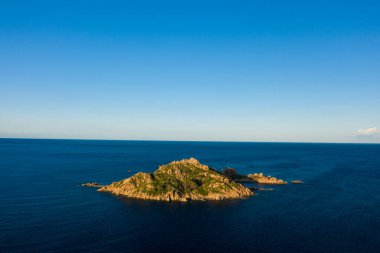 Yeşil bitki örtüsüyle kaplı kayalık bir ada, geniş ve berrak bir gökyüzünün altında, Lotzorai, Sardunya yakınlarındaki sakin mavi Akdeniz sularıyla çevrilidir. Sıcak güneş ışığı manzaranın doğal dokularını ve hatlarını vurgular.