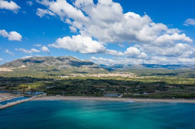 Parlak turkuaz dalgalar geniş kumlu bir kumsalla buluşur, bereketli kırsal ve dramatik dağ sıraları ile Orosei, Sardinya yakınlarında dağınık bulutlarla dolu bir gökyüzünün altında.