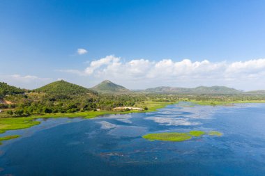 Brateak Gölü 'nün geniş mavi suları Kamboçya' daki Kampong Chhnang 'da açık bir gökyüzünün altında yuvarlanan yeşil tepelere ve uzak tepelere doğru uzanıyor. Sudaki bitki örtüleri ve dağınık ağaçlar, gün ışığında sakin göl kıyıları boyunca uzanır..