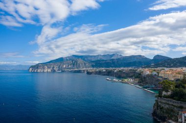 Sorrento 'nun dramatik uçurumları ve renkli binaları derin mavi Akdeniz sularına tepeden bakar ve gökyüzünde geniş bulut oluşumları oluşur. Yat ve tekneler marina boyunca demirli, engebeli dağların arkasına kurulmuş.