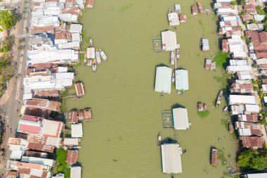 Vietnam, Chau Doc 'ta yüzen bir köyün doğrudan manzarasında geniş, yeşilimsi bir nehrin her iki tarafında sıralanmış yapılar ve teneke çatılı binalar görülüyor. Küçük tekneler ve yüzen yapılar, eşsiz bir yerleşim yeri olan suyu gösteriyor.