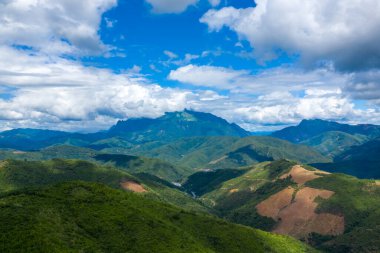 Geniş yeşil tepeler ve orman yamaçları kuzey Laos 'ta dağınık bulutlarla dolu canlı mavi bir gökyüzünün altında dramatik bir dağ sırasına doğru uzanıyor. Manzarada ekilmiş toprak, yuvarlanan arazi parçaları ve uçsuz bucaksız doğal bir his yer almaktadır.