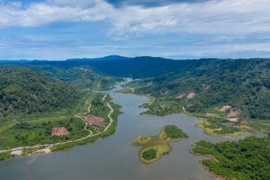 Yoğun ormanlık tepeler ve bulutlu bir gökyüzünün altındaki temiz toprak parçalarının arasında dönen bir nehrin havadan görünüşü. Çevrede dolambaçlı su, yeşil bitki örtüsü ve uzak bir kırsal bölgede doğal kıvrımlar bulunur..