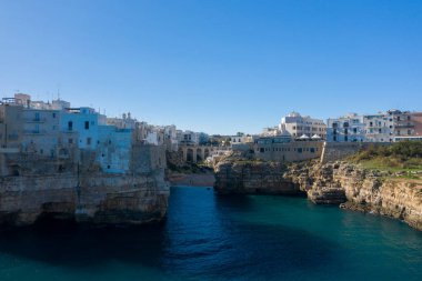Polignano a Mare, Apulia, İtalya 'daki turkuaz su üzerindeki dramatik kireçtaşı kayalıkları boyunca beyaz ve taş binalar kümelenir. Kıyı sahnesinde canlı güneş ışığı, desenli kaya ve açık mavi gökyüzü yer almaktadır..