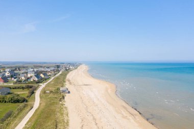 Sweeping aerial perspective of Utah Beach with pale sand, turquoise water, and a line of houses along grassy dunes under a clear blue sky. Bright daylight creates a spacious, tranquil seaside mood.