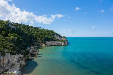 İtalya 'nın Peschici kenti yakınlarındaki Adriyatik kıyısında berrak turkuaz suların üzerinde yoğun yeşil çam ormanlarıyla kaplı dik kireçtaşı uçurumları yükseliyor. Sahne, parlak güneş ışığıyla aydınlatılıyor. Canlı, mavi bir gökyüzünün altında, dağınık bulutlarla.