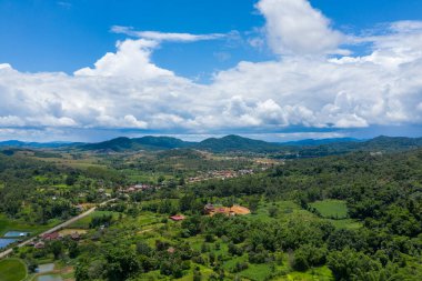 Bereketli yeşil tepeler ve yoğun bitki örtüsü arasında kurulu bir kırsal köy perspektifi, dalgalanan bulutlarla dolu canlı mavi gökyüzünün altındaki katmanlı dağlara kadar uzanıyor. Manzara, geniş kırsal ve doğal bölgeleri yakalıyor.