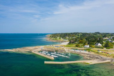 Taştan rıhtımlar, canlı sahil kulübeleri ve turkuaz suları olan, yeşil bir sahil köyüne ve parlak bir gökyüzünün altındaki açık denize bakan, kompakt bir marinanın havadan görüntüsü..