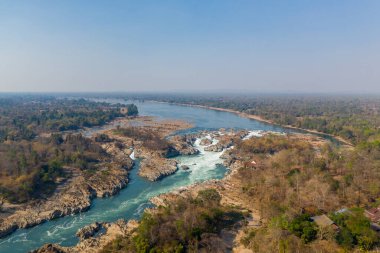Laos 'un güneyindeki Mekong Nehri' ndeki Chutes de Khone 'un hava perspektifi, temiz mavi gökyüzünün altında kurak bir orman ve geniş bir nehir ovası arasında engebeli kayaların üzerinde akan güçlü beyaz suyu gösteriyor. Geniş manzara dramatikliği vurguluyor.