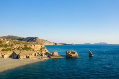 Triopetra plajı boyunca uzanan sakin mavi sulardan, engebeli uçurumlar ve berrak bir gökyüzünün altındaki uzak dağlarla ayırt edici kumtaşı yığınları yükselir. Sahne huzurlu, geniş ve doğal olarak çarpıcı..