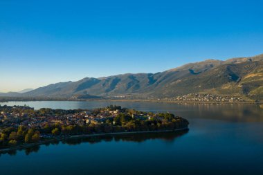 Ioannina Gölü 'nün sakin sularıyla çevrili tarihi Ioannina yarımadasını, arka planda yükselen engebeli Pindus dağlarıyla sakin bir hava manzarası yakalar. Sahnede kırmızı kiremitli çatılar, yemyeşil yemyeşil ve geniş mavi gökyüzü yer alıyor.