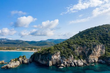 Yunanistan 'ın Epirus kentindeki Karavostasi' de, turkuaz kıyı şeridi boyunca uzanan engebeli kayalık kayalıklardan yemyeşil yamaçlar yükseliyor. Parlak gün ışığı ve dağınık bulutlar Akdeniz manzarasını ve doğal dokuları vurgular.
