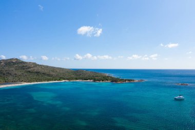 Sardunya 'nın Spiaggia la Salinedda yakınlarındaki berrak turkuaz sularda yüzen bir katamaranın havadan görünüşü, yemyeşil tepeler ve küçük bulutlarla benekli parlak mavi bir gökyüzü..