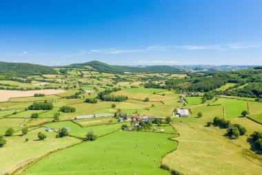 Morvan bölgesindeki yuvarlak tepelerin arasına kurulmuş güneşli arazi, yeşil çayırlar ve dağınık çiftlik binalarının hava manzarası. Manzara berrak mavi bir gökyüzü altında canlı, açık ve huzurlu..