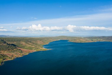 Lac de Salagou 'nun yuvarlanan yeşil tepeler ve kırmızı toprakla çevrili derin mavi sularıyla geniş hava manzarası. Dağınık bulutlarla genişleyen gökyüzü, açık alan ve sükunet hissine katkıda bulunur..