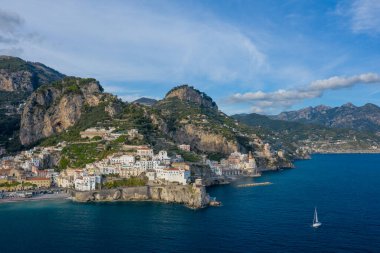 Tek bir yelkenli, beyazlatılmış binaların ve İtalya 'nın Amalfi kentindeki engebeli kayalıkların yanında derin mavi sularda süzülüyor. Sahne dramatik Akdeniz kıyılarını, teraslı yamaçları ve berrak gökyüzünün altındaki uzak dağları yakalıyor..