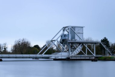 Çağdaş beyaz çelik asma köprü, çıplak ağaçlar ve bulutlu gökyüzünün altında yumuşak gün ışığıyla çevrili sakin bir nehir boyunca uzanır ve sakin ve endüstriyel bir atmosfer yaratır..
