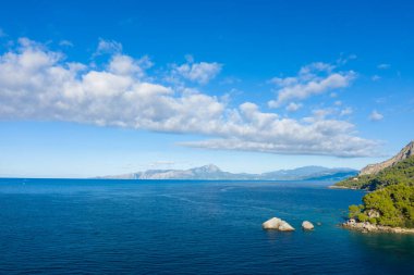 Uçsuz bucaksız mavi deniz manzarası kayalık kıyı şeridi ve dağılmış bulutlarla dolu parlak bir gökyüzünün altındaki uzak dağ sıraları. Güneş ışığı, kıyı manzarasının doğal güzelliğini ve huzurunu vurgular..