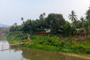 Luang Prabang, Laos 'taki yemyeşil tropikal bitki örtüsünün ve palmiye ağaçlarının altındaki sakin nehir kıyısını kırsal bir bambu köprüsü geçer. Sahnede, yumuşak, puslu bir gökyüzünün altında yoğun yaprakların arasına yerleştirilmiş küçük beyaz bir ev ve ahşap teras yer alıyor..