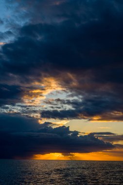 Alacakaranlıkta Adriyatik Denizi 'nin üzerinde toplanan koyu mavi ve altın bulutlar, gökyüzünde çarpıcı ışık şeritleri oluşturarak sakin suları yansıtıyor. Sahne, gölgelerin çarpıcı bir etkileşimini ve açık havada ılık günbatımı tonlarını yakalar.