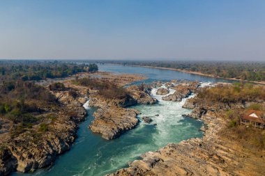 Güney Laos 'taki Mekong Nehri' ndeki Khone Phapheng Şelalesi 'nin geniş hava manzarası. Kuru ormanlarla çevrili engebeli kayalar ve berrak mavi gökyüzünün altında geniş bir nehir manzarası üzerinde dalgalı beyaz su akıntıları..