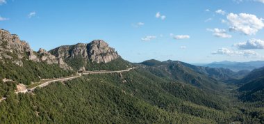 Sardunya 'daki sık yeşil ormanların üzerinde engebeli bir dağ sırtının kenarındaki yüksek yol geniş manzaralı ve açık mavi gökyüzünün altında kayalık tepeler..