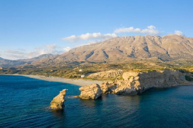 Girit 'in Triopetra plajındaki derin mavi Akdeniz sularının üzerinde uzanan kumtaşı kayalıkları ve deniz yığınları engebeli dağlar ve berrak bir gökyüzünün altındaki mesafeye uzanan güneşli bir kıyı şeridi..